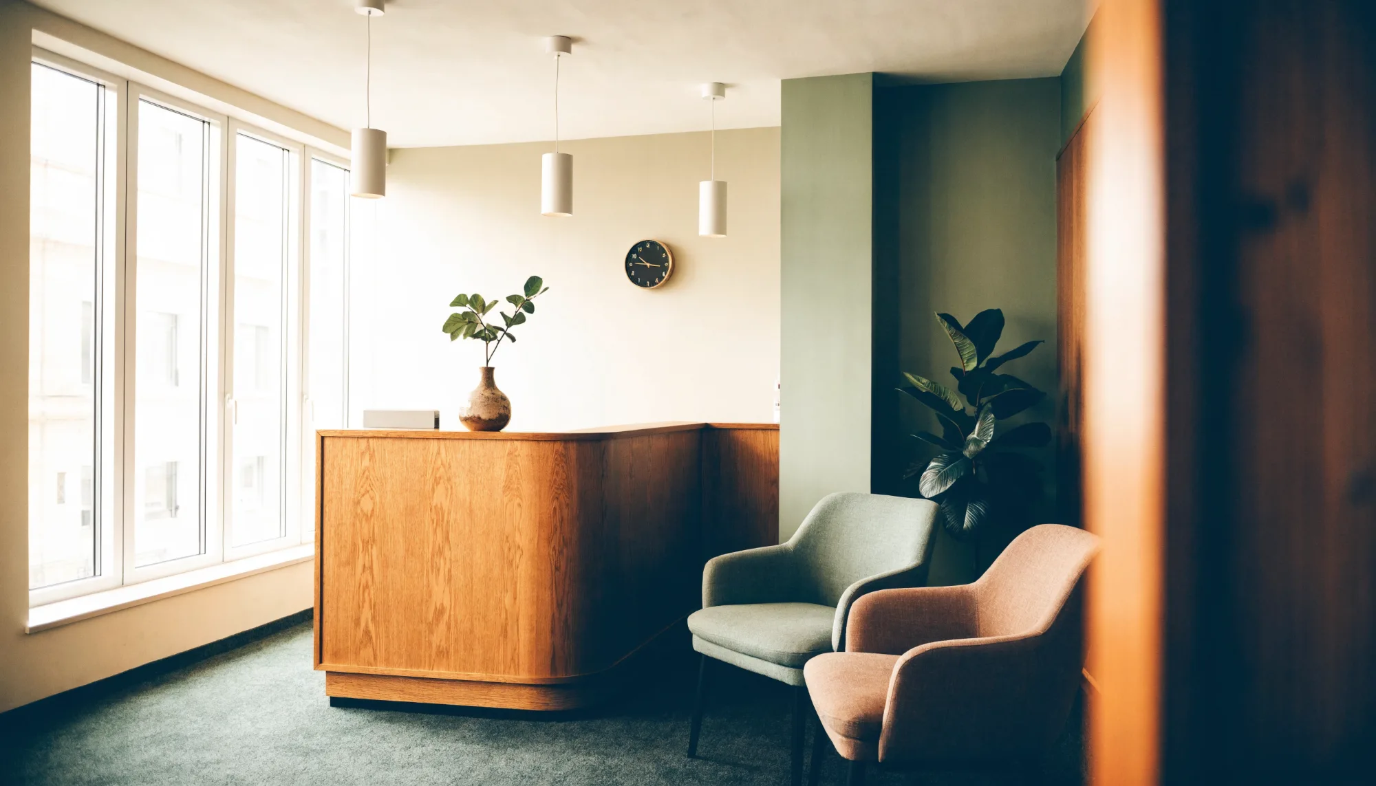 The reception and waiting area at Alder House: an oak desk with an alder sprig, a sage chair and a clay chair, and a clock on the wall.