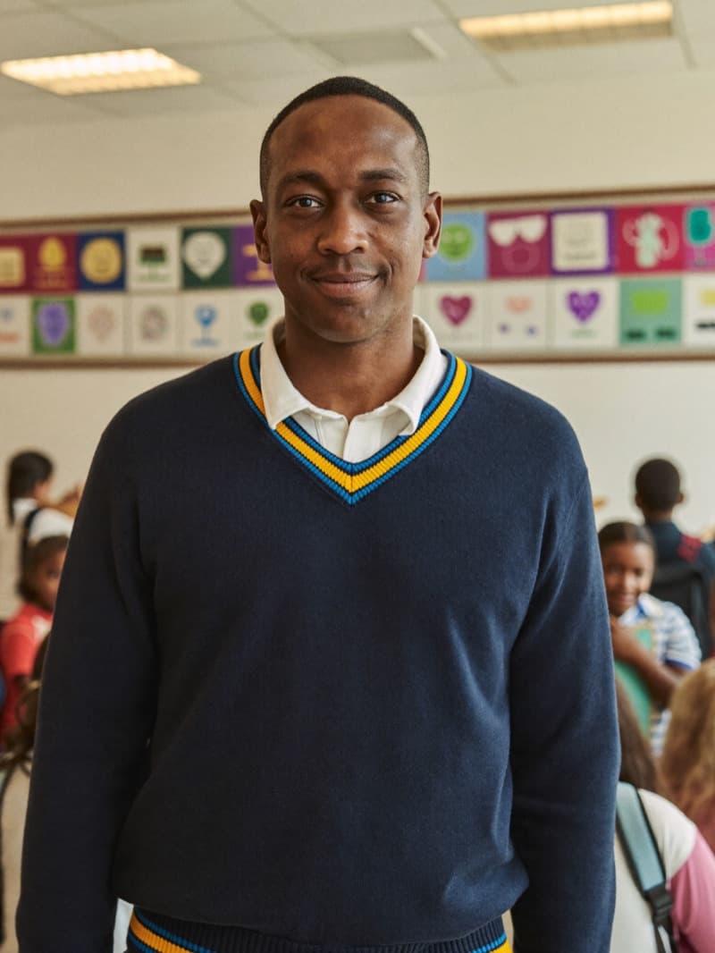 Mr David Okonkwo, Deputy Head and Designated Safeguarding Lead, smiling in the school hall in a navy school jumper, children in the background.