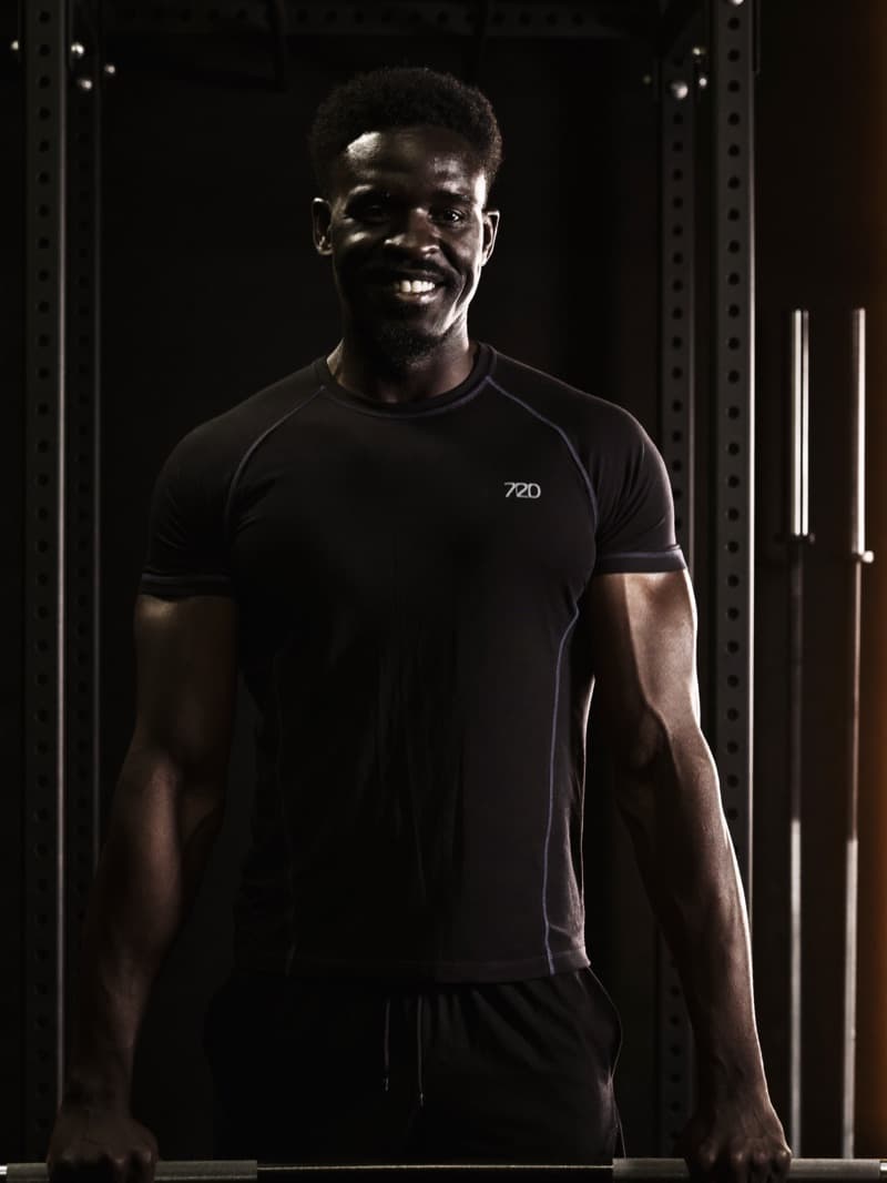 Danny Aluko, Strength Coach, arms folded on the gym floor in warm low light