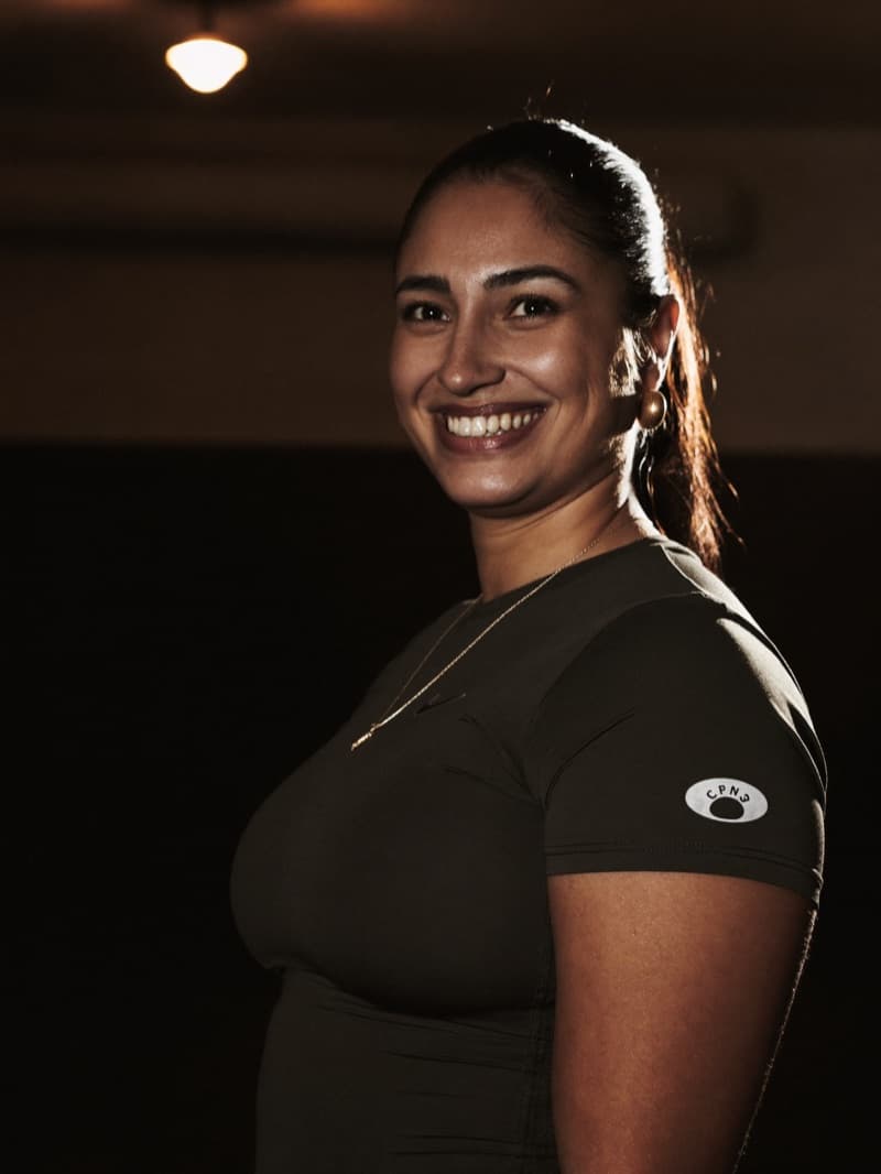 Priya Sharma, Foundations Coach, smiling by the gym door under warm low light